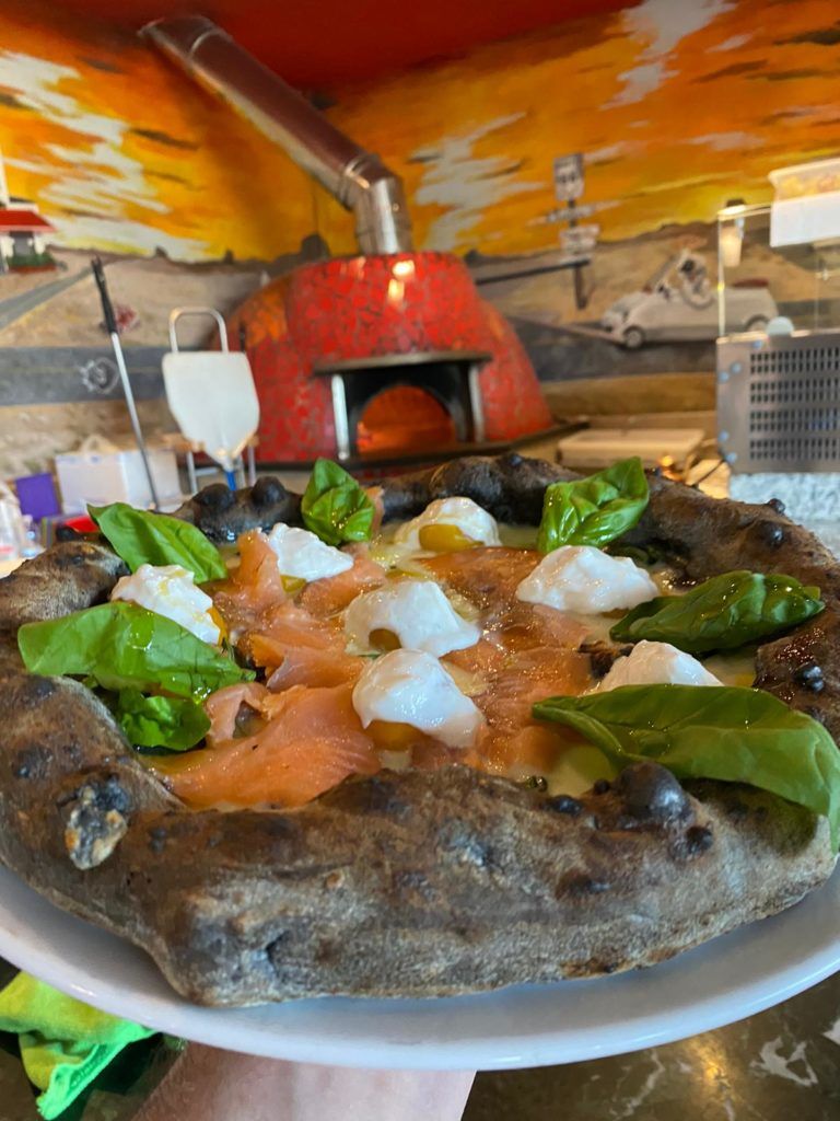 Pizza appena sfornata con salmone, stracciatella e basilico, tenuta davanti al forno a legna rivestito di mosaico rosso